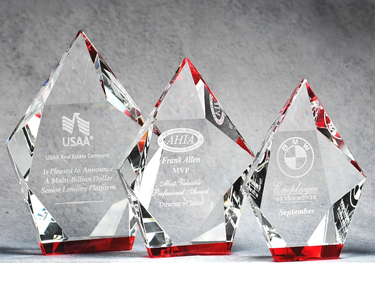 Three pentagon-shaped crystal awards with engraved text and logos on a gray background