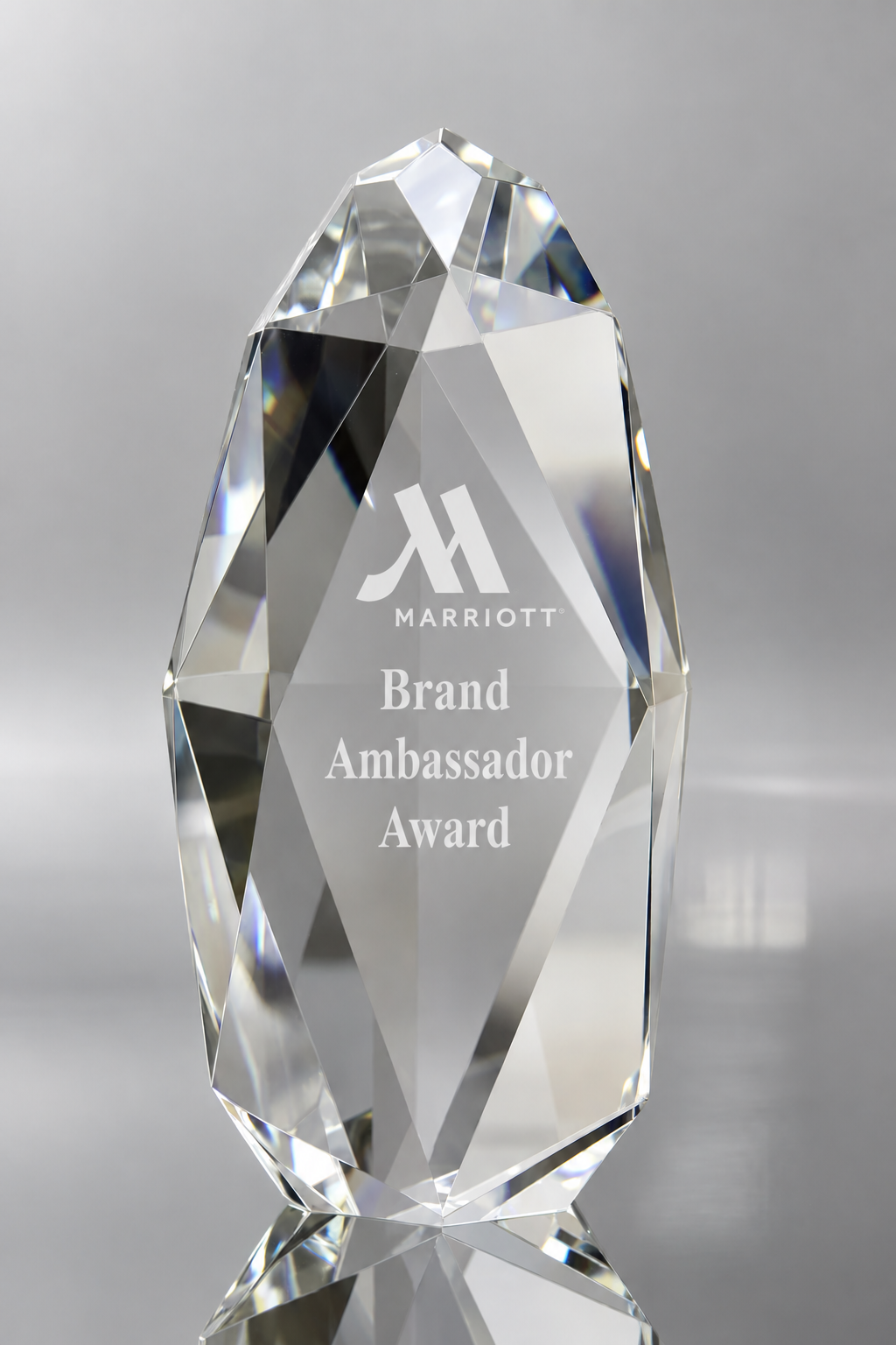 A faceted clear crystal award trophy engraved with the Marriott logo and the text 'Brand Ambassador Award,' shown on a reflective gray surface against a soft gray background.