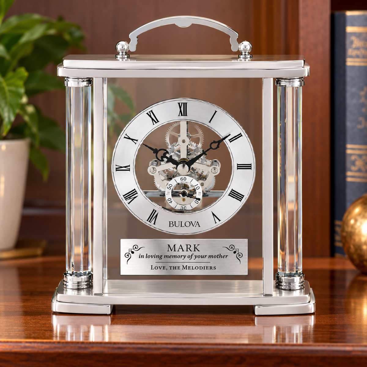 Bulova Adrienne silver mantel clock with glass columns, exposed movement, and personalized engraving plate on a wooden shelf.