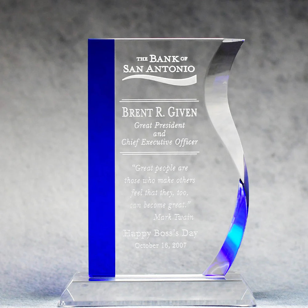 A blue and clear crystal glass award with engraved text, sitting on a optic crystal base.