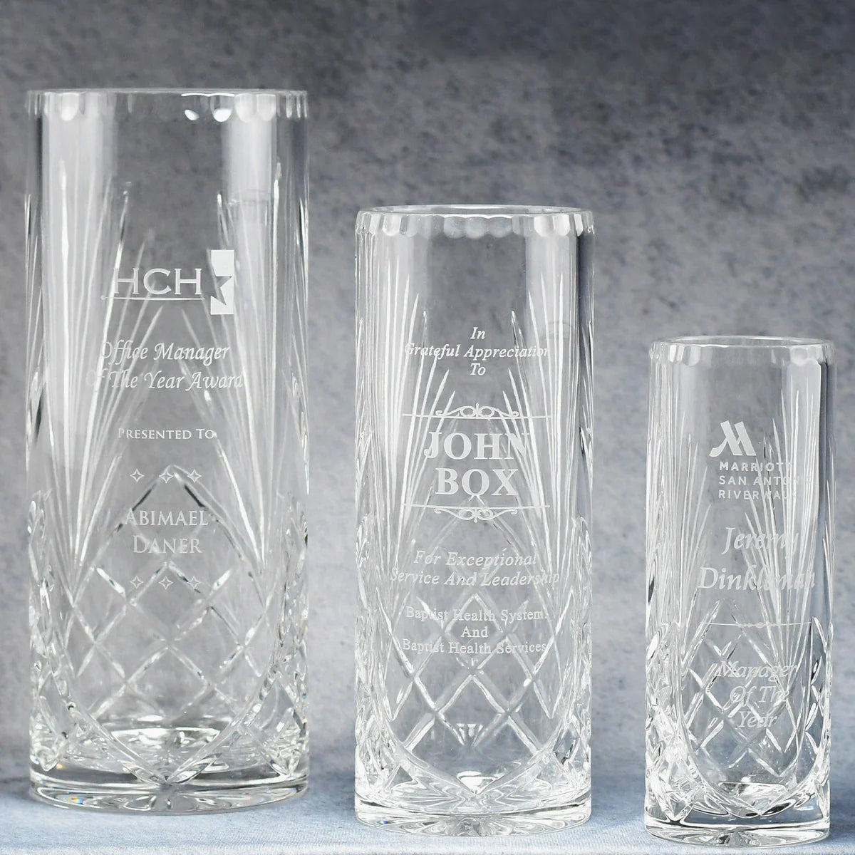 Three clear crystal cylinder vases of different sizes with engraved text and images on them, displayed against a grey background.