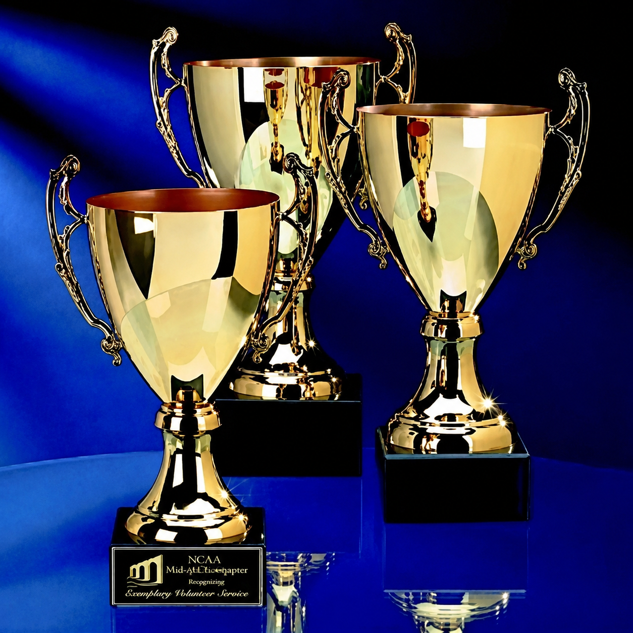 Three polished gold trophy cups with black bases displayed on a reflective blue surface against a dark blue background. The front trophy has a black plaque