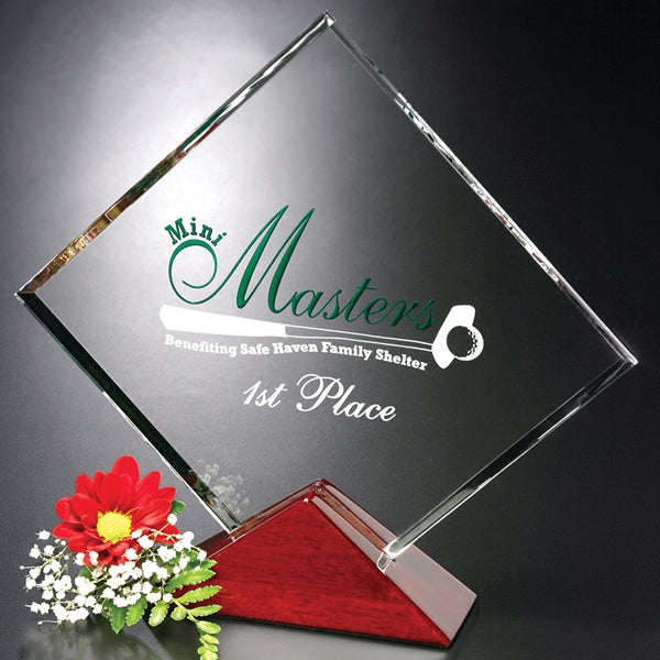 A square optical crystal award with engraved text on a piano finished base, accompanied by a red and white flower arrangement.