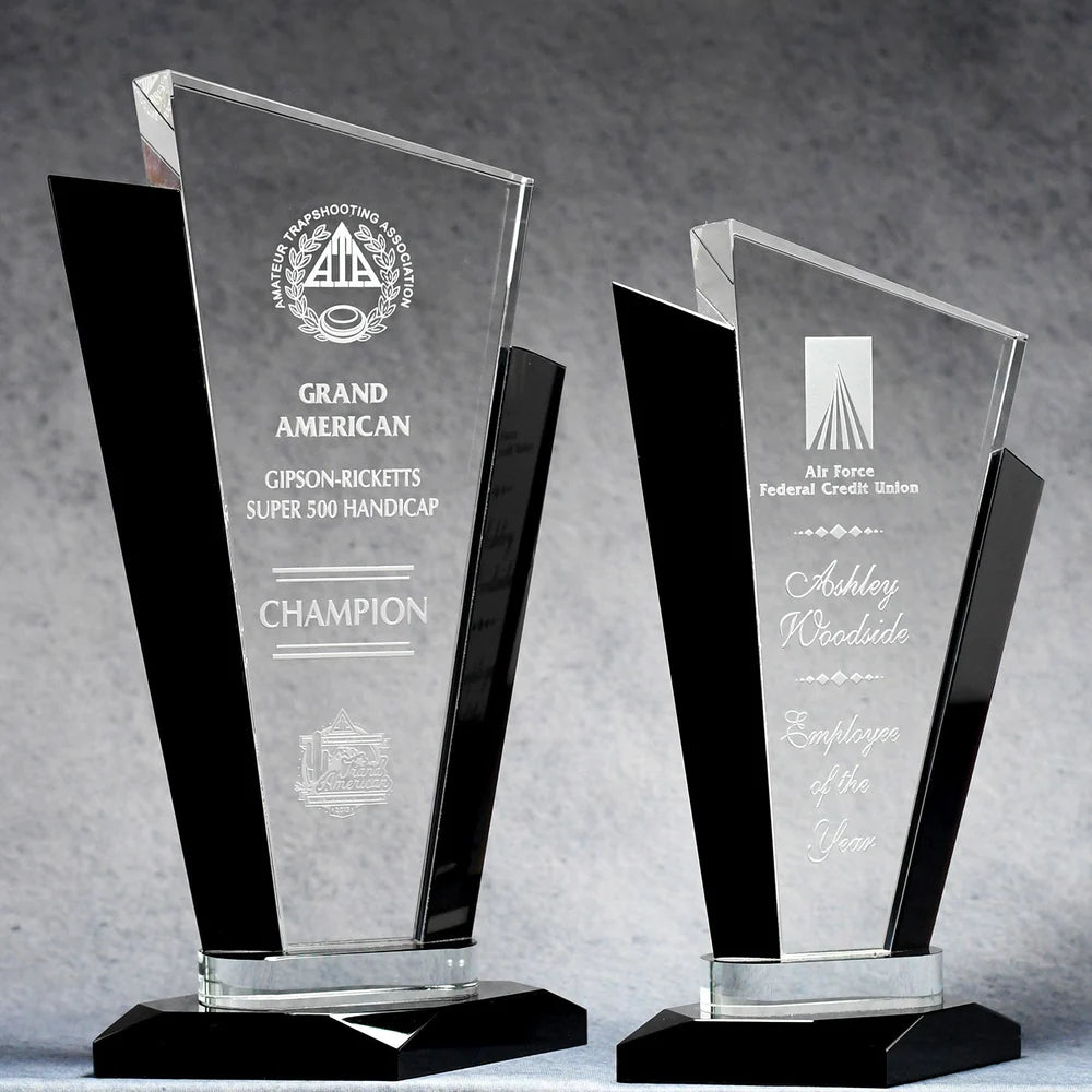 Two crystal tuxedo trophy awards of different sizes with black accents and etched text, placed on a grey surface.