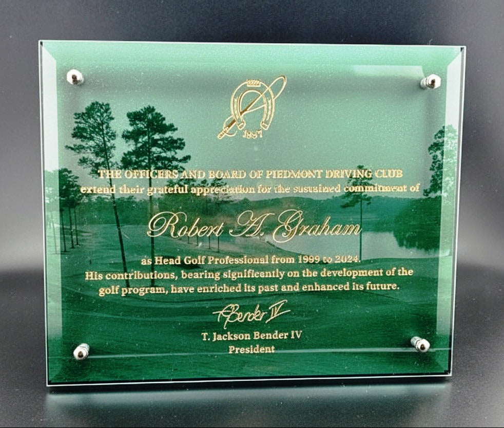 glass plaque with gold text and a logo, displayed on a dark surface with a blurred background of trees.
